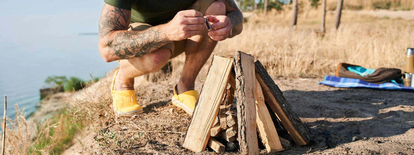 Man crouching down in shorts behind a wood stack that he is about to light up with matches