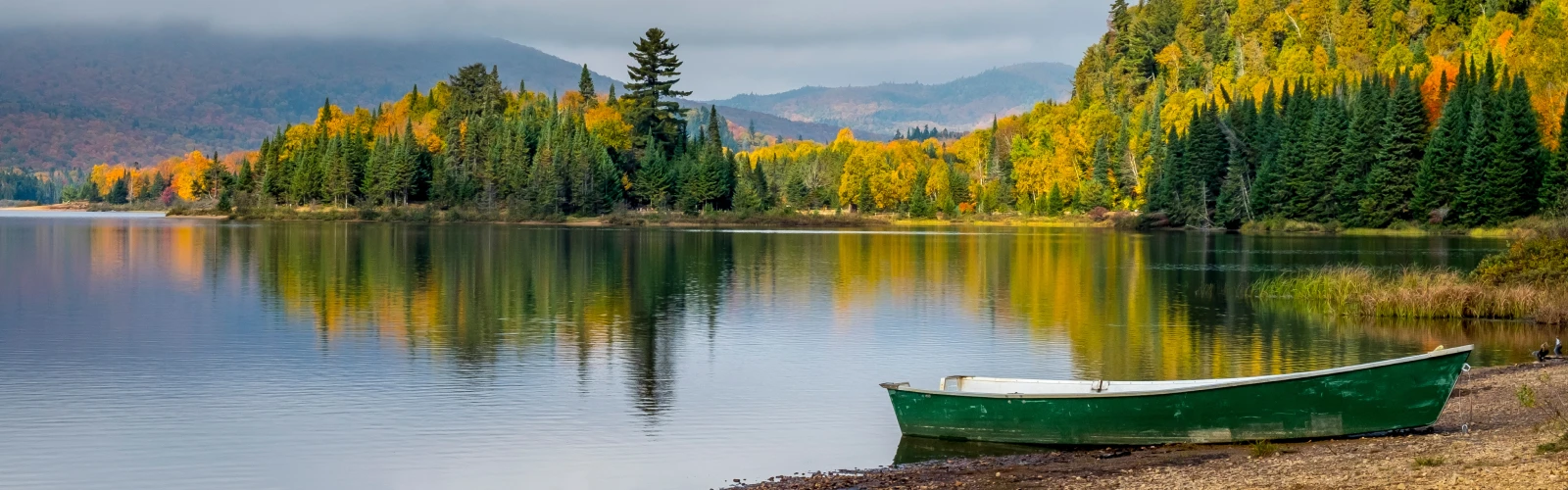 A row boat sits on the shore of a lake with fall trees in the distance